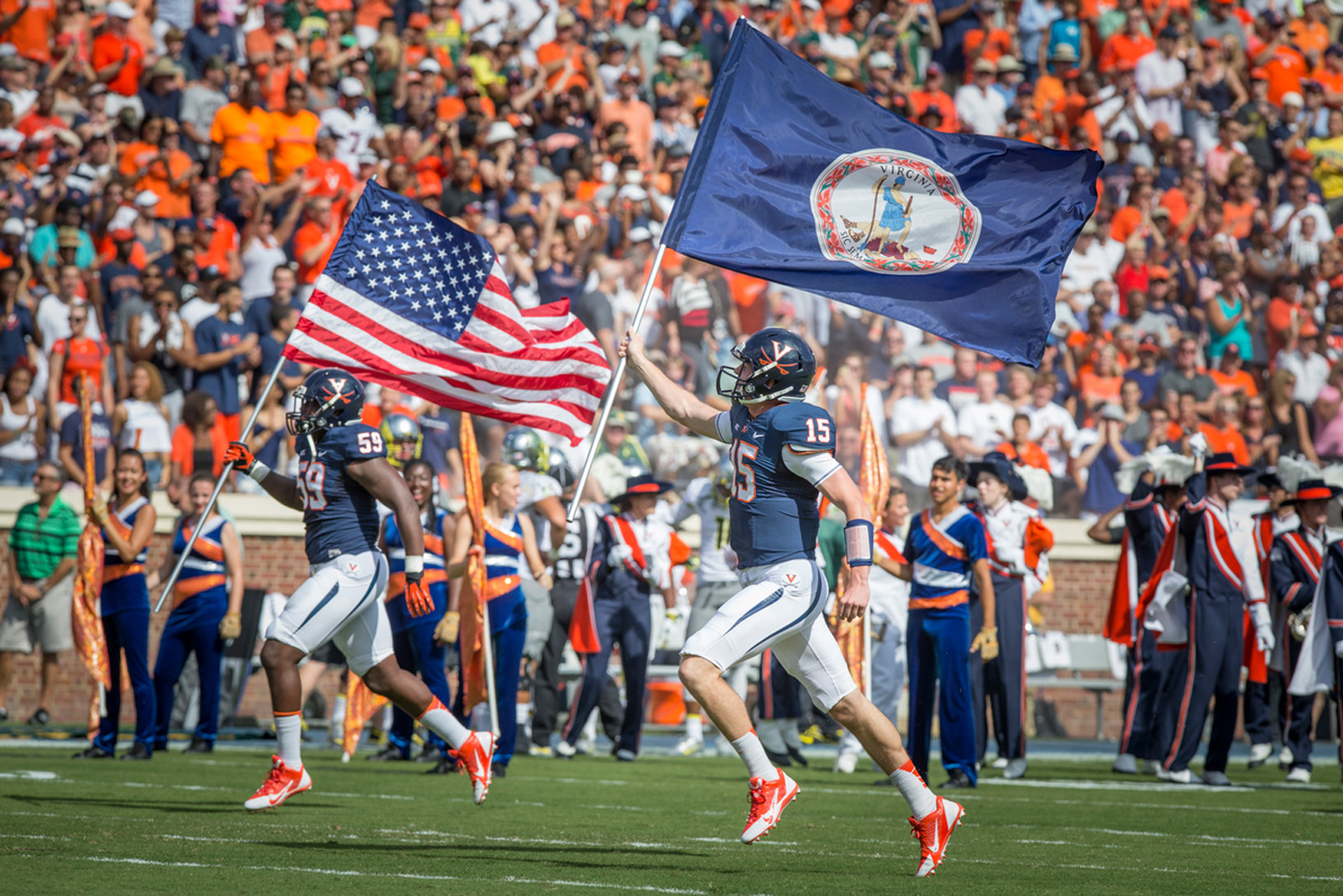 A portrait of two UVA football players running in the stadium with the USA and Virginia flags, with a cheering crowd in the background.